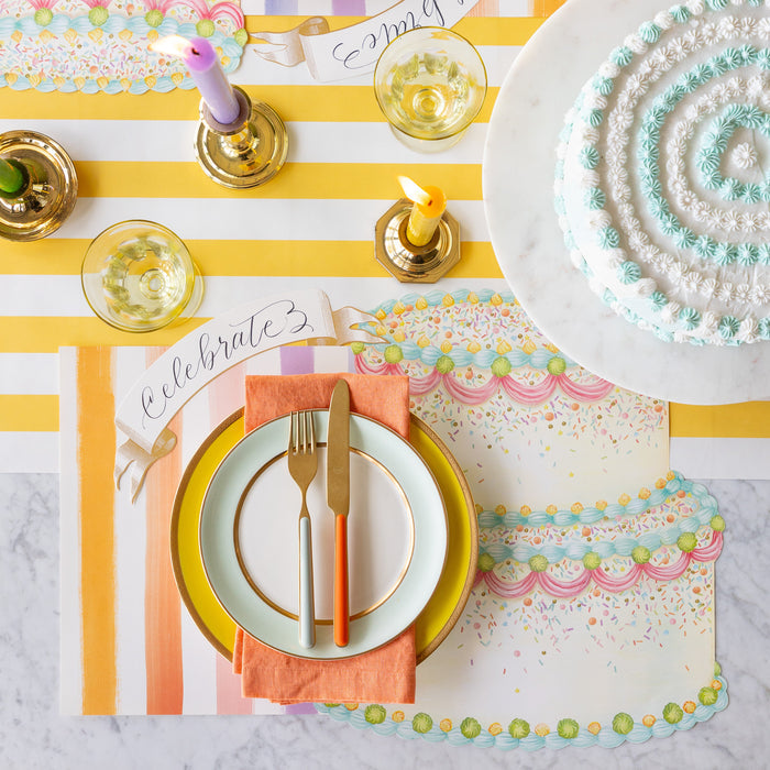 A colorful table setting featuring the Die-cut Birthday Cake Placemat overlapping the Sorbet Painted Stripe Placemat, colorful dinnerware and Classic Gold Banner Table Accent on the Marigold Classic Stripe Runner.