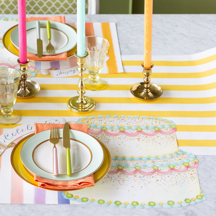 A colorful table setting featuring the Die-cut Birthday Cake Placemat overlapping the Sorbet Painted Stripe Placemat, colorful dinnerware and Classic Gold Banner Table Accent on the Marigold Classic Stripe Runner.