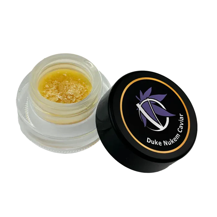 Angled top-down view of an open jar of Duke Nukem Caviar showing golden, crumbly concentrate inside and the black branded lid beside it.
