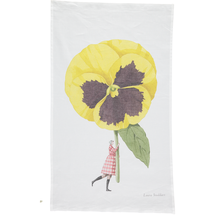 A white tea towel featuring a whimsical illustration of a small woman holding a gigantic yelllow pansy by the stem. Art by Laura Stoddart.
