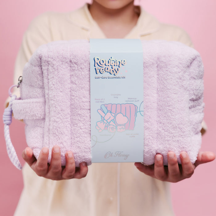 A girl in lemon pajamas holds a light purple plush cosmetic bag with a matching wrist strap. The pouch has a central paper label that reads 'Routine Ready' and 'Cosmetic Essentials Kit' with a cute illustration.