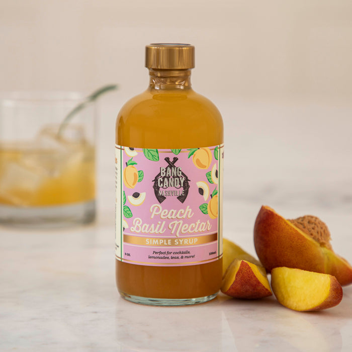 A bottle of Hester & Cook Peach Basil Nectar Syrup next to peach slices with a drink garnished with basil in the background.