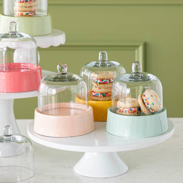 A close-up of a light blue, light pink, and yellow Petit Sweet Cake Dome atop a white cake stand, some with treats inside.