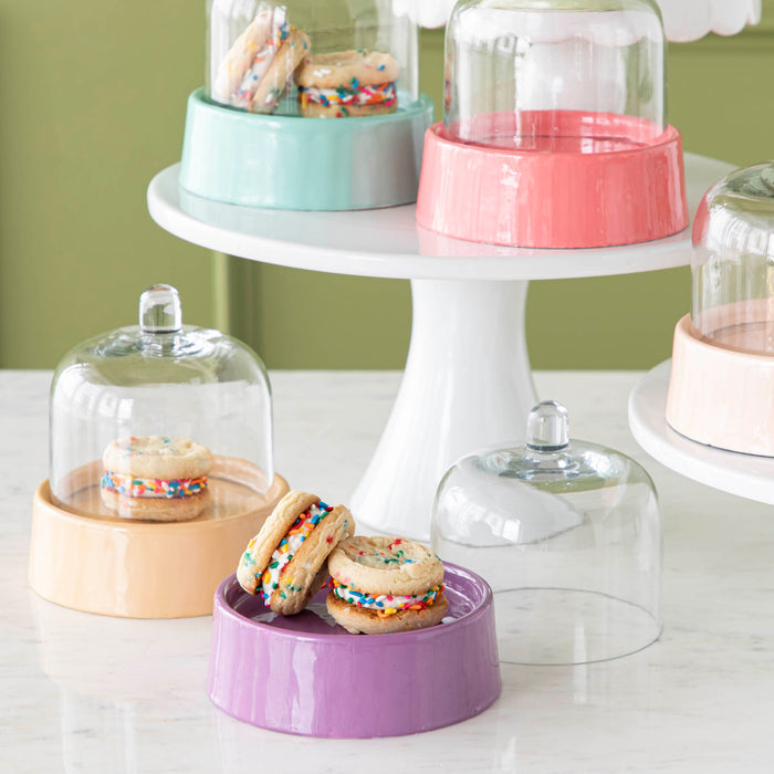 Two Petite Sweet Cake Domes in the colors sherbert and violet, one with the top removed, sitting atop a marble surface with treats inside.